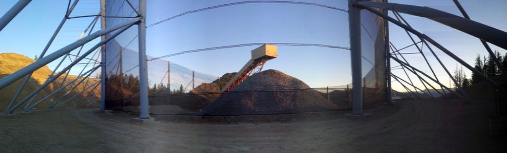 A large pile of gravel sits beneath a conveyor belt, surrounded by a metal framework and netting