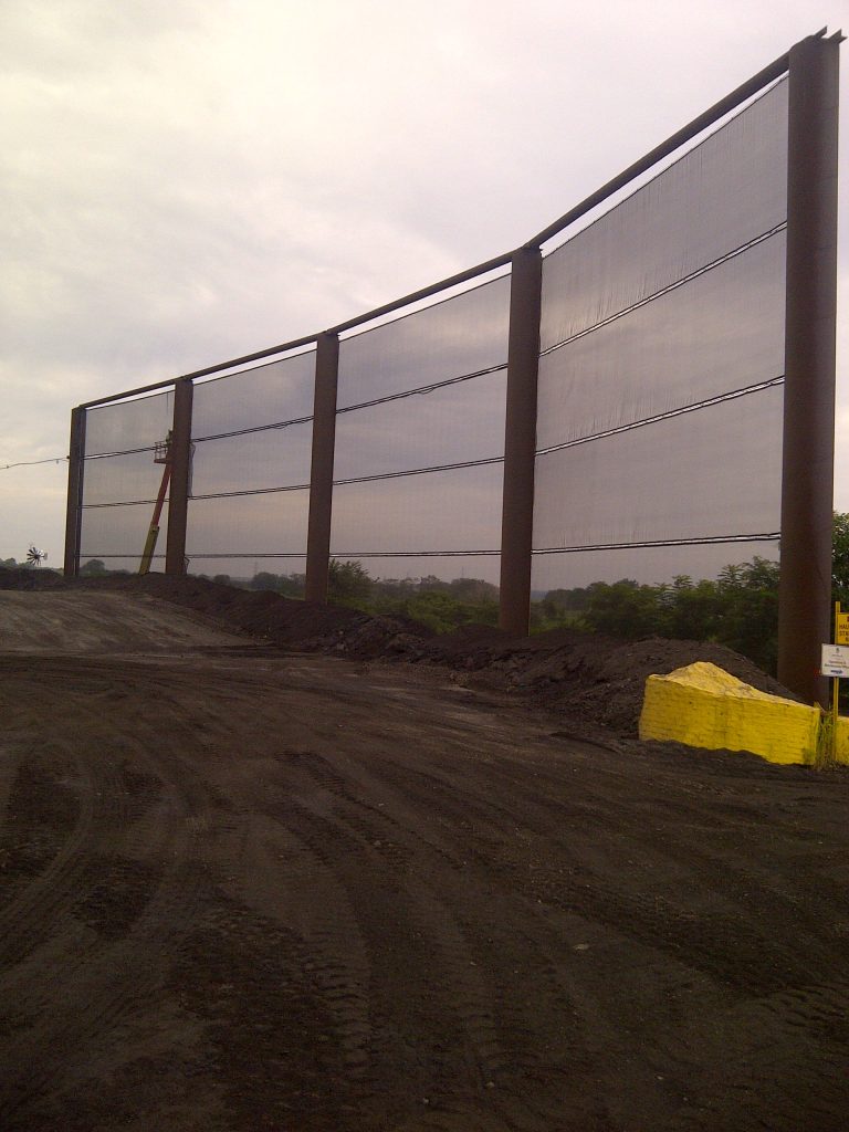 A large industrial wind fence with metal posts and translucent panels stands on a dirt site