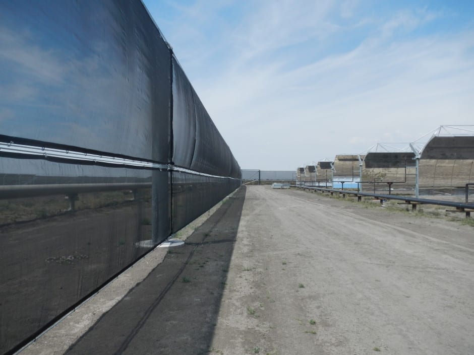 A long black fence runs alongside a dirt road with large structures in the background