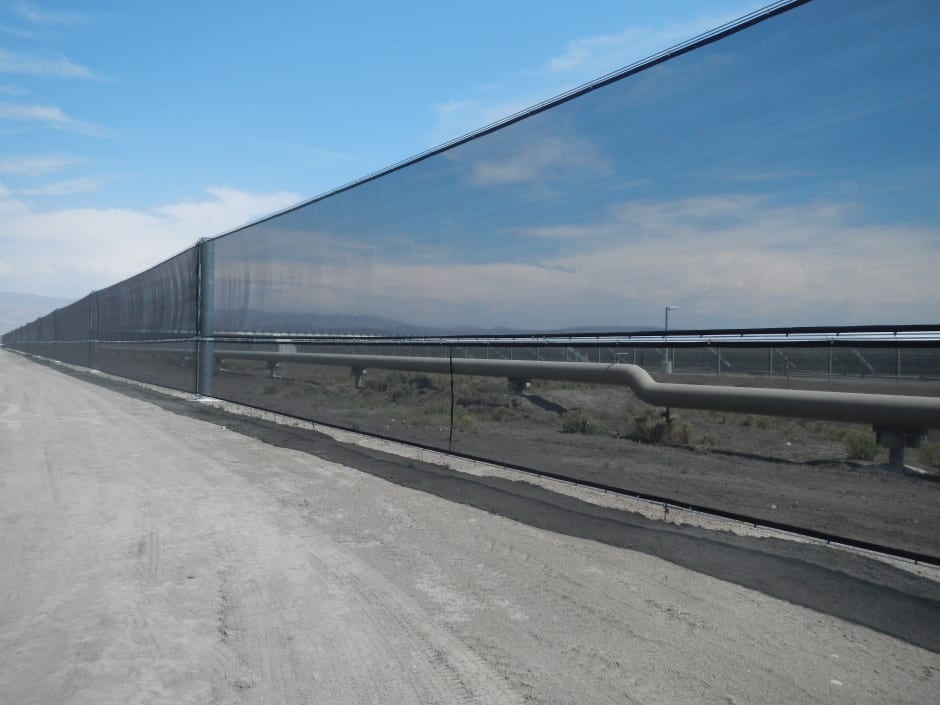 A long, reflective fence stretches alongside a dirt road under a clear sky