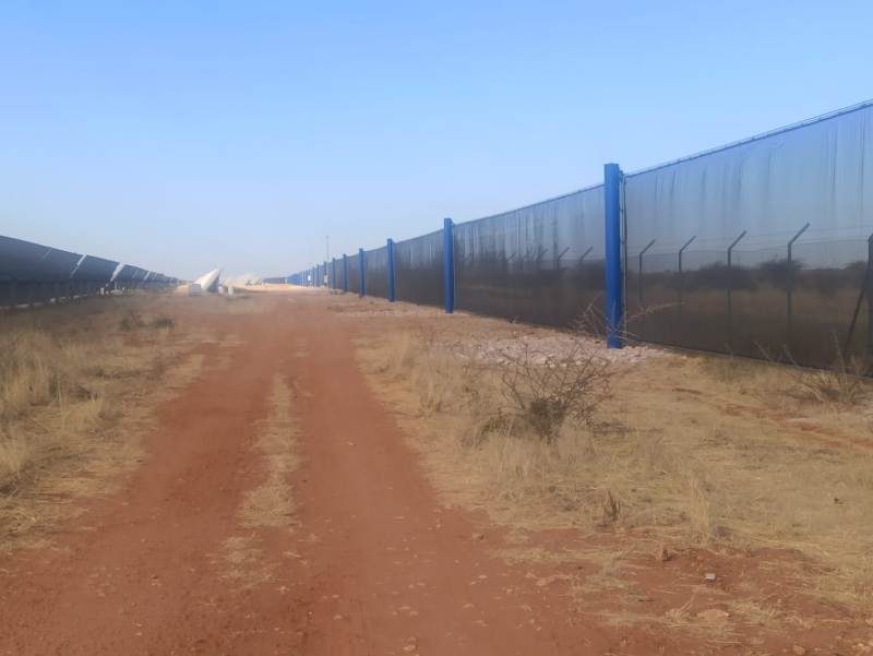 A dirt path runs between solar panels and a long blue wind fence
