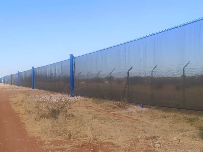 A long blue and black wind fence stretches across a dry, grassy area under a clear sky