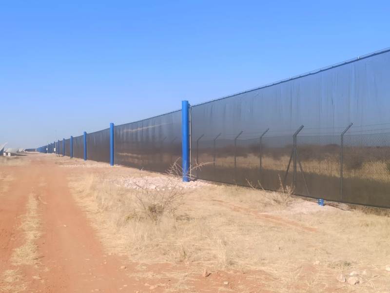 A long blue and black fence stretches across a dry, grassy landscape under a clear sky