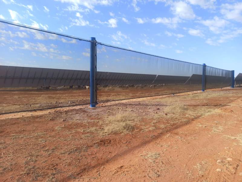 A long, reflective wind fence stretches across a dry, reddish landscape under a partly cloudy sky