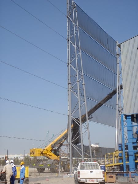 A tall industrial wind fence with workers and machinery at the base under a clear sky