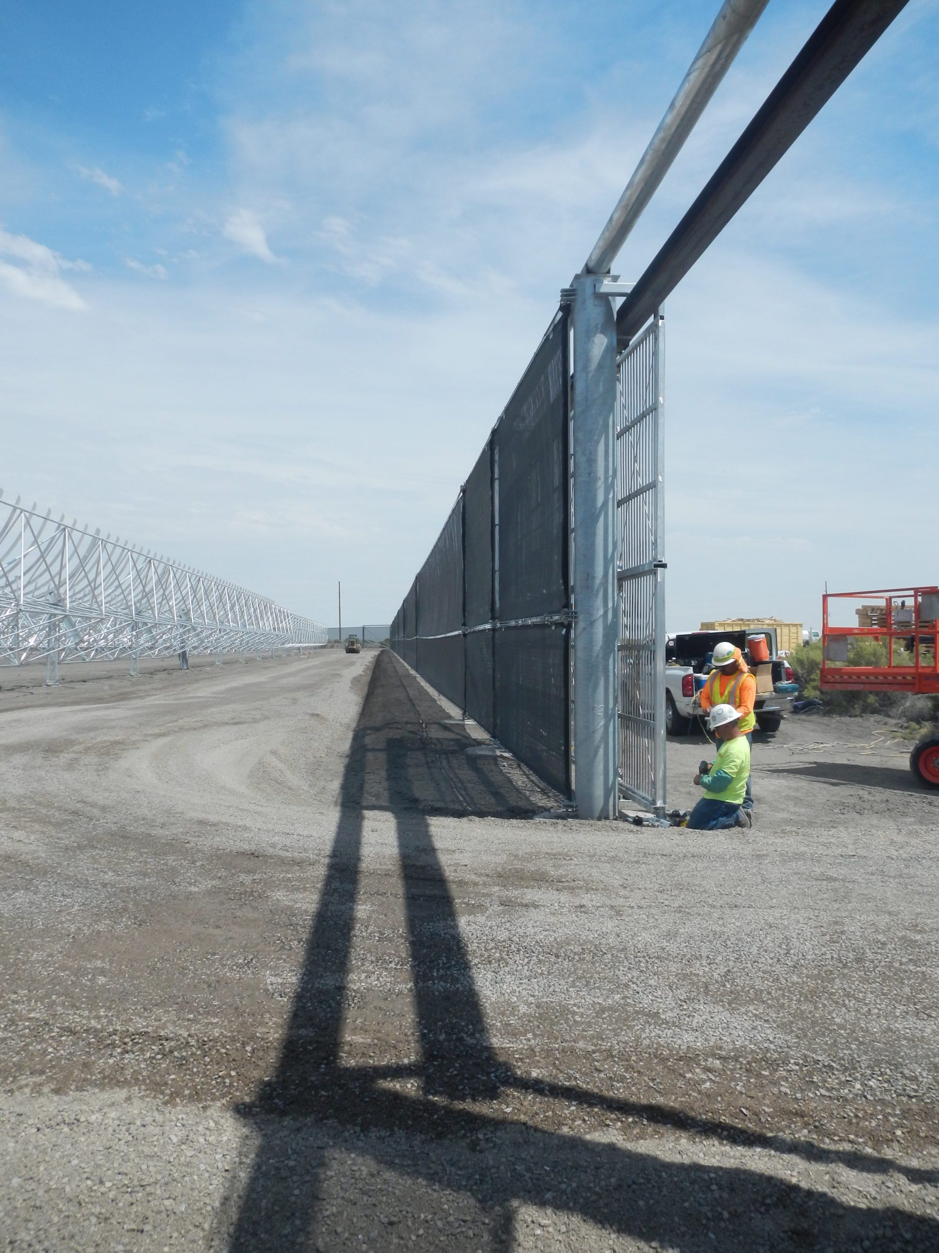 Workers in safety gear install a tall metal fence along a dirt road, with construction equipment and vehicles nearby under a clear blue sky.