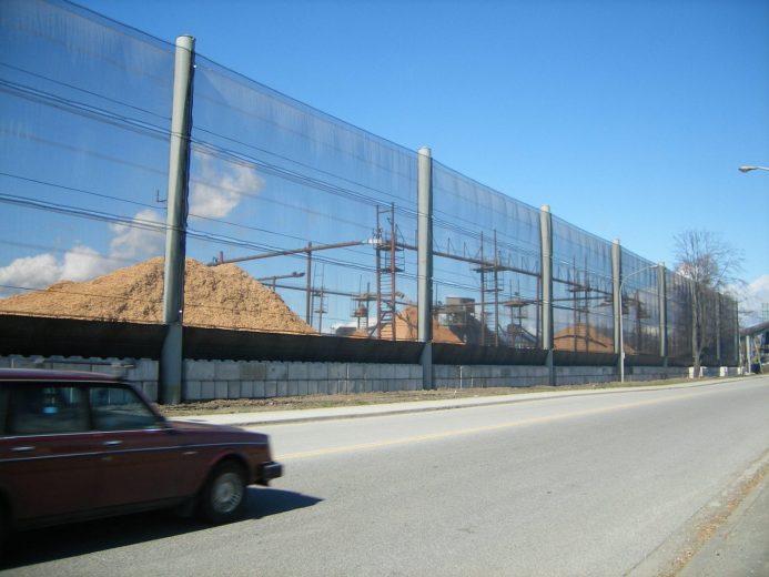 A long, transparent fence runs alongside a road, with piles of wood chips behind it