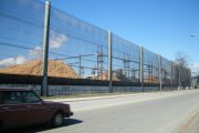 A long, transparent fence runs alongside a road, with piles of wood chips behind it