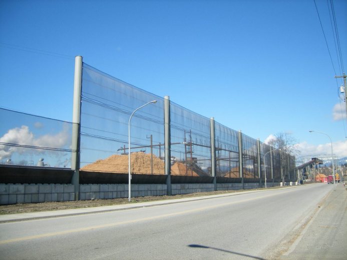 A long, transparent wind fence runs alongside a road, with piles of material visible behind it