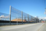 A long, transparent wind fence runs alongside a road, with piles of material visible behind it