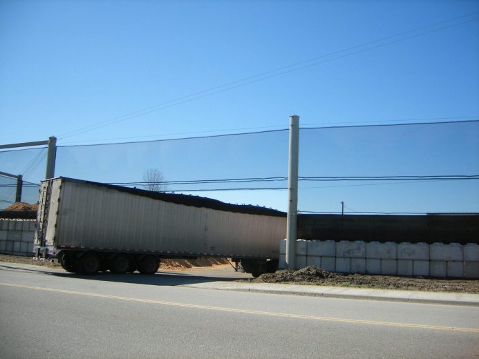 A semi-truck trailer is positioned under power lines beside a concrete barrier