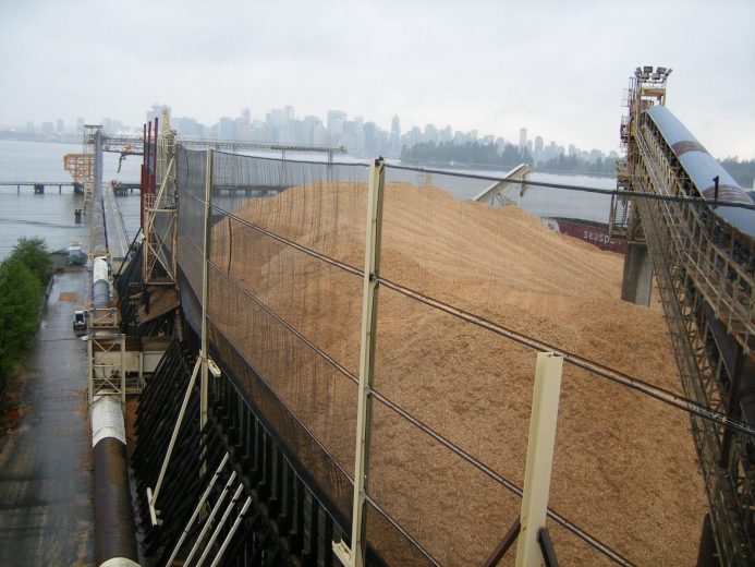 A large pile of grain is covered by a wind fence near a waterfront city