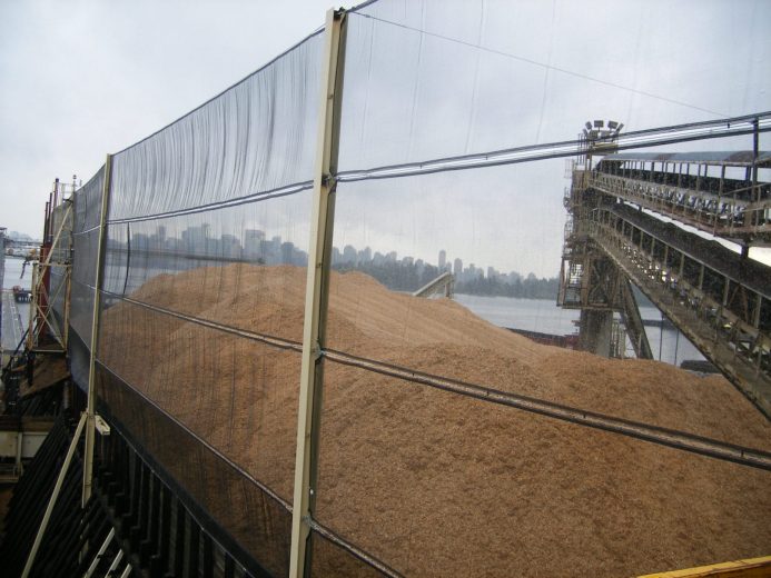 A large pile of wood chips is covered by a transparent wind fence on a dock
