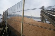A large pile of wood chips is covered by a transparent wind fence on a dock