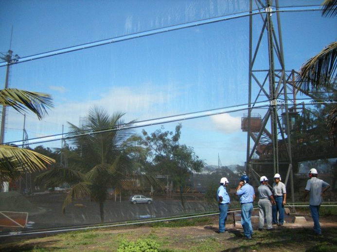 Four construction workers wearing hard hats stand near a large metal structure and netting
