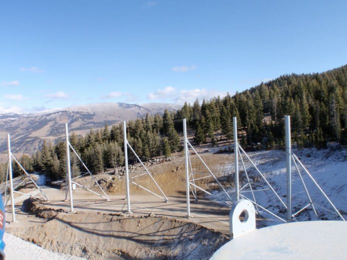 Metal poles and supports installed on a sloped, snowy ground with trees and mountains in the background