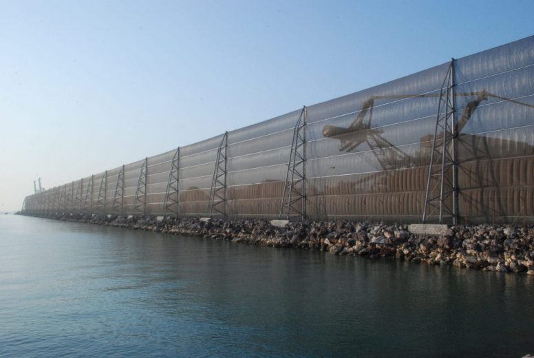 A long, transparent wind fence stretches along a rocky shoreline with a crane visible behind it