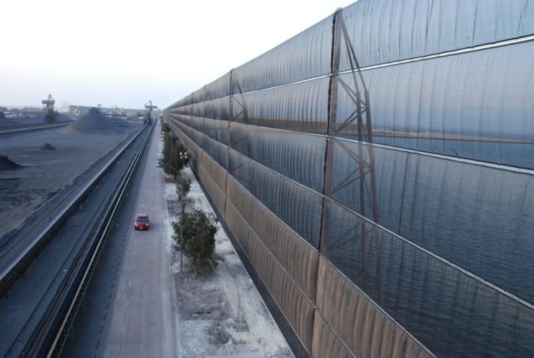 A long industrial wind fence stretches alongside a road with a red car driving past