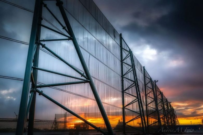 A large industrial fence structure stands against a dramatic sunset sky