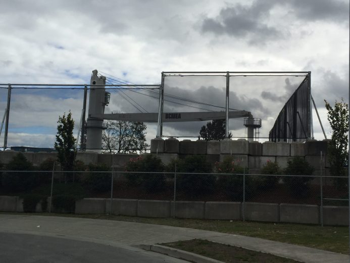A large industrial crane stands behind a chain-link fence and concrete barriers