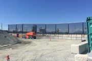 Construction site with orange machinery, gravel, concrete barriers, and a black mesh fence
