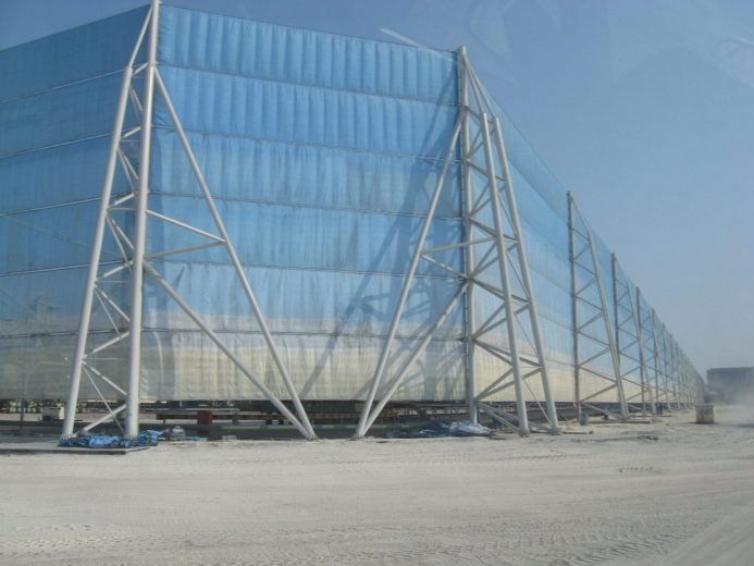 A large, blue, translucent structure supported by a white metal framework stands on a dusty, unpaved surface