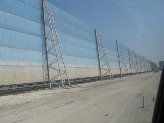 A long, light blue wind fence stretches across a sandy area under a clear sky