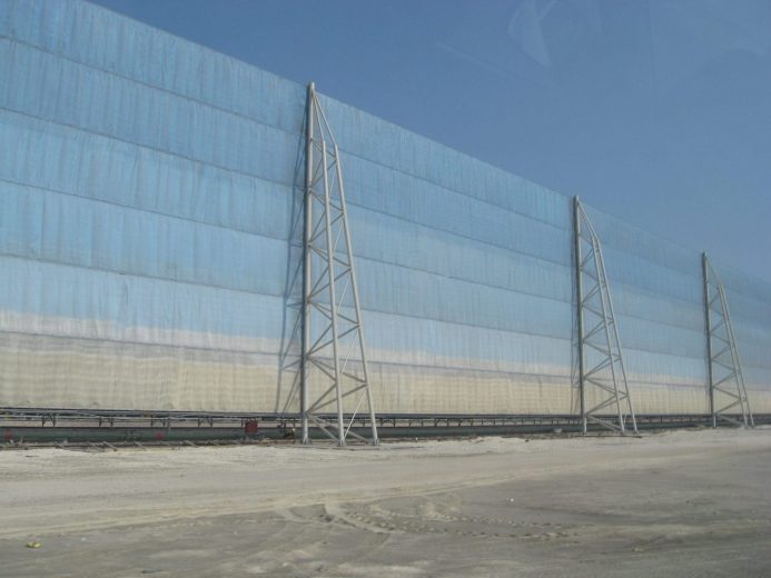 A large, light blue wind fence supported by metal towers stands against a clear sky