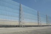 A large, light blue wind fence supported by metal towers stands against a clear sky