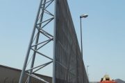 A tall, triangular metal structure stands next to a road with an orange JLG lift nearby