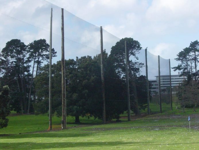Tall poles with netting stretch across a grassy area with trees and a building in the background