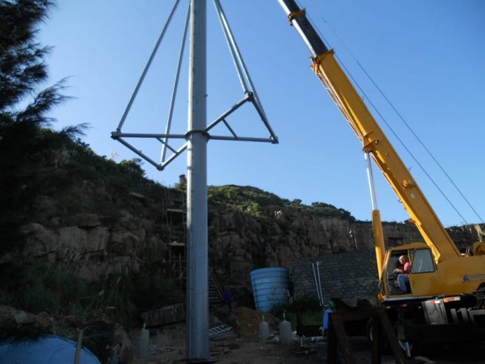 A crane lifts a tall metal pole with triangular supports against a clear blue sky