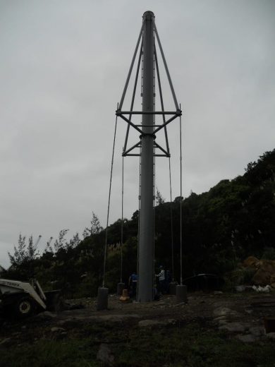 A tall metal pole with guy wires and a ladder, surrounded by construction equipment and workers