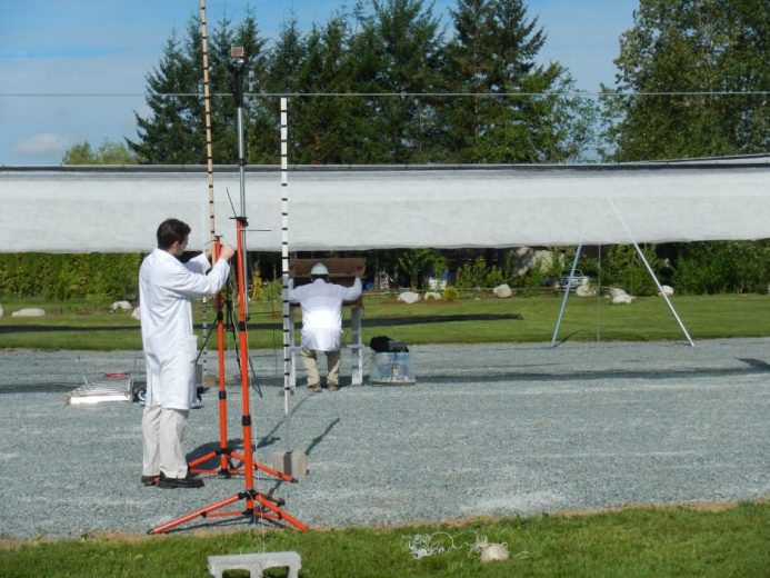 Two scientists in white lab coats work with equipment under a large white canopy outdoors