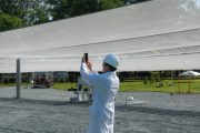 A person in a lab coat and hard hat takes a photo under a large white canopy