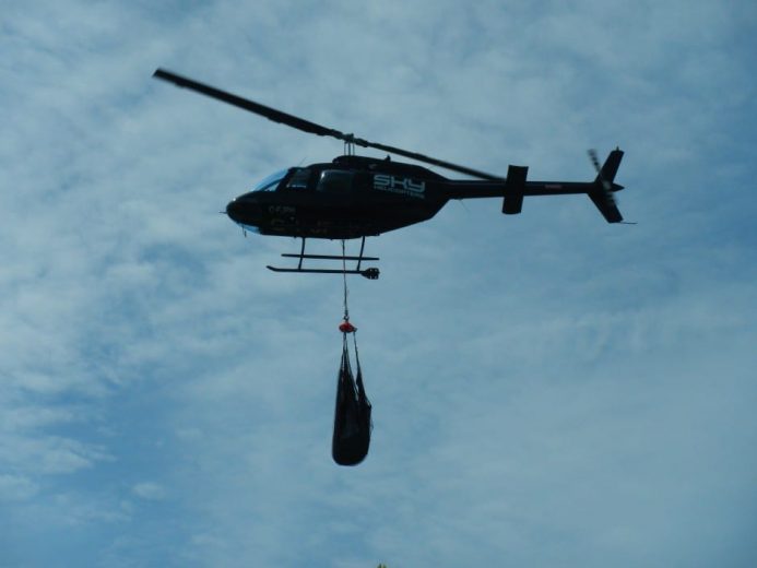 A helicopter lifts a large, dark object suspended by cables against a cloudy sky