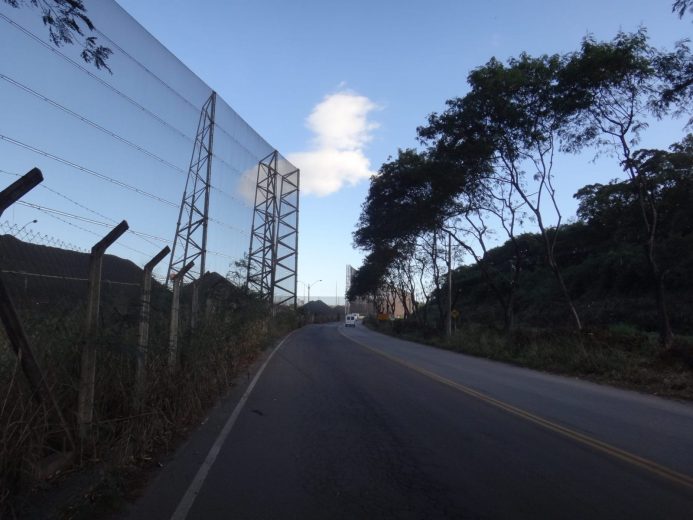 A road curves to the right with a tall fence and trees on either side