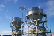 Two large industrial silos with yellow railings and a tall light pole under a clear blue sky