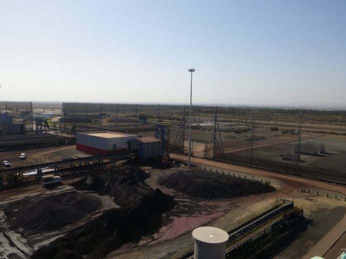 An industrial site with piles of dark material, buildings, and fencing under a clear sky
