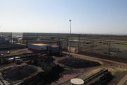 An industrial site with piles of dark material, buildings, and fencing under a clear sky