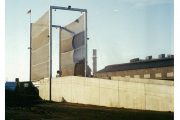 A large industrial wind fence stands against a clear sky, with a concrete wall and industrial buildings in the background
