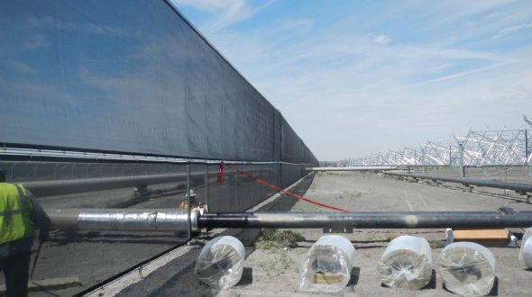 A long black wind fence stretches across a construction site with pipes and wrapped materials in the foreground