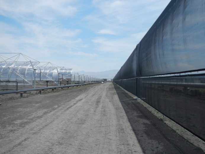 A long dirt road stretches between a row of metal structures and a tall black fence