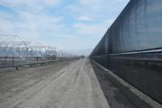 A long dirt road stretches between a row of metal structures and a tall black fence