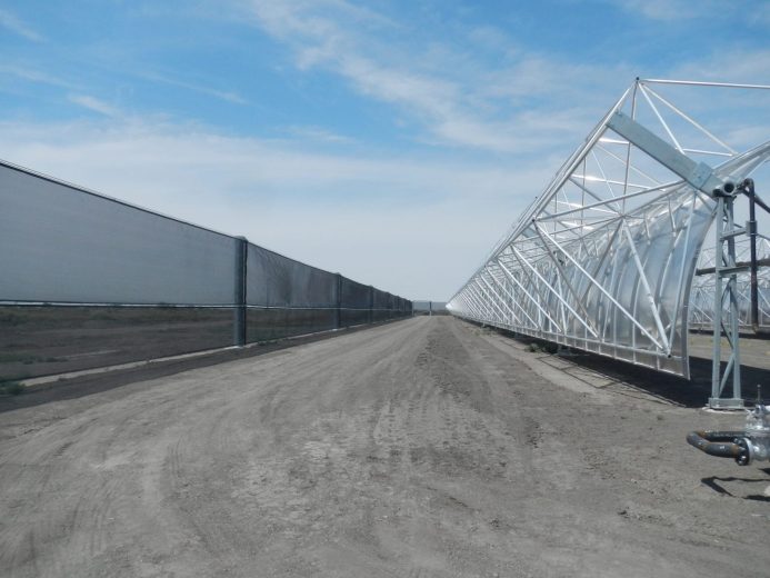 A long dirt road stretches between a tall wind fence and a metal structure