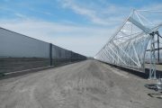 A long dirt road stretches between a tall wind fence and a metal structure