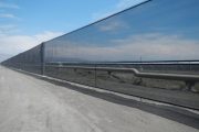 A long black mesh fence stretches alongside a dirt road under a clear sky