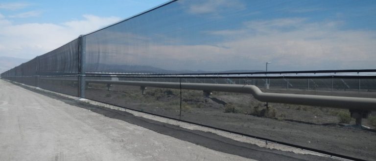 A long black mesh fence stretches alongside a dirt road under a clear sky