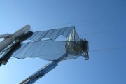 A person in a crane basket adjusts a translucent wind fence against a clear blue sky