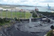 A coastal industrial area with a large wind fence, conveyor belt, and shipping cranes